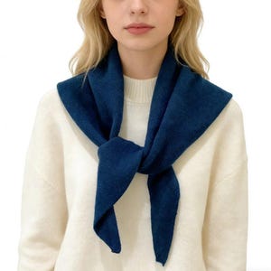 May include: A dark teal scarf is tied around the neck, creating a layered look. The scarf is made of a soft, textured material and is worn over a white sweater. The scarf's ends are knotted in the front.