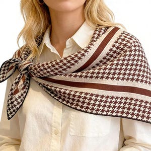 May include: A brown and white houndstooth patterned scarf tied around the neck. The scarf has a dark brown border and stripes. The scarf is draped over a white collared shirt. The scarf is made of a soft material.