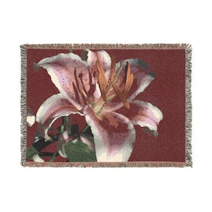 May include: A woven throw blanket featuring a large lily flower in shades of pink, white, and brown against a burgundy background. The blanket has a fringed edge.