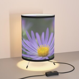 Może przedstawiać: Cylindryczna lampa stołowa z kwiatowym wzorem. Klosz lampy przedstawia zbliżenie fioletowego kwiatu z żółtym środkiem. Lampa ma czarne wykończenie, białe wnętrze i podstawę statywu. Lampa jest podłączona do gniazdka elektrycznego.