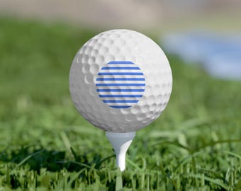 May include: A white golf ball with a blue and white striped design, resting on a white tee in a field of green grass. The golf ball has a dimpled surface and a circular graphic.