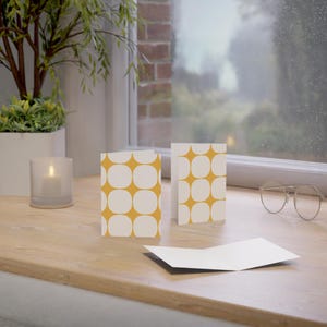 May include: Greeting cards with a retro design. The cards feature a pattern of white rounded squares on a mustard yellow background. One card is open, and two are standing upright. A lit candle and eyeglasses are also visible.
