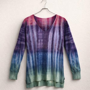 Puede incluir: Un suéter de cuello en V de manga larga con un diseño tie-dye. Los colores cambian de rosa y morado en la parte superior, a azul y morado en el medio, y a verde en la parte inferior. El suéter está colgado en una percha de metal.