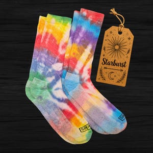 May include: A pair of ribbed crew socks featuring a vibrant tie-dye pattern in shades of red, yellow, blue, and purple. A brown rectangular paper tag is attached with a string, displaying the text Starburst, a sun icon, and a crescent moon.