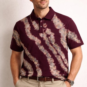 May include: Burgundy polo shirt with a tie-dye pattern in shades of gray and beige. The shirt has a pocket on the chest and a classic collar. The model is wearing khaki pants and a brown leather belt.
