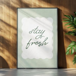 Puede incluir: Una impresión artística enmarcada con las palabras "stay fresh" en escritura verde. El texto está sobre una forma blanca con bordes festoneados sobre un fondo verde pálido. El marco es negro y la impresión se muestra contra una pared de madera.