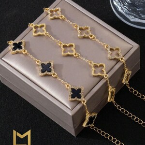 Op de afbeelding: Twee goudkleurige schakelarmbanden met klaverbladmotieven. De ene armband heeft zwarte emaille klaverbladcentra, terwijl de andere open klaverbladframes heeft, beide versierd met kleine heldere steentjes langs de randen.