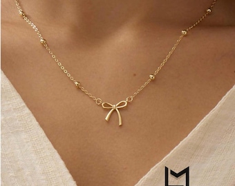 May include: A delicate gold-colored necklace featuring a small bow pendant. The necklace has a fine chain with evenly spaced small beads. The bow pendant is centered, and the necklace is displayed against a neutral background.