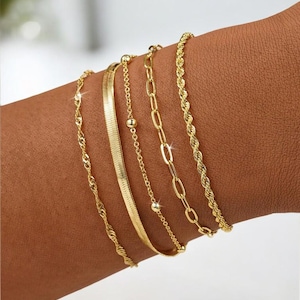 Puede incluir: Conjunto de cinco pulseras doradas con diseños variados. Incluyen una cadena trenzada, un diseño plano en espiga, una cadena delicada con pequeñas cuentas, una cadena con eslabones ovalados y una cadena de estilo cuerda. Las pulseras se muestran en una muñeca.