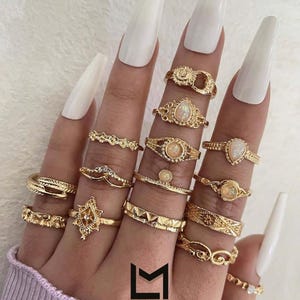 May include: A hand displaying a collection of gold rings with various designs, including filigree, geometric patterns, and gemstone accents. The rings are worn on multiple fingers, showcasing their different styles. The image includes the text "L'ÉMET MARCHE".