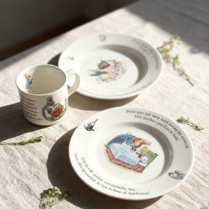 May include: A white ceramic mug and plates featuring illustrations of a rabbit in a blue coat. The plates have text around the rim. The mug and plates are on a beige tablecloth, with sunlight casting shadows.