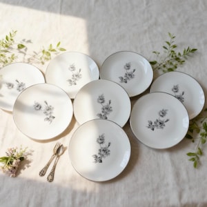 May include: A set of eight white porcelain plates with a delicate floral design in the center and a silver rim. The plates are arranged on a white tablecloth, with sprigs of greenery and silverware.
