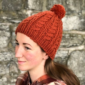 May include: A knitted beanie hat in a burnt orange color with a cable knit pattern and a fluffy pom pom on top.
