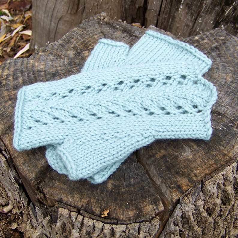 May include: A pair of light blue knitted fingerless gloves with a lacy pattern. The gloves are laid out on a tree stump.