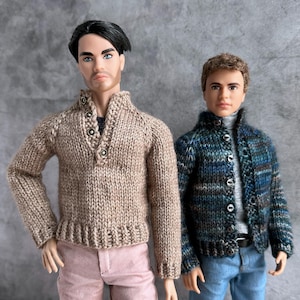 以下が含まれることがあります： 2体の人形がニットセーターを着ています。1体はボタン付きの襟とピンクのズボンが付いた薄茶色のセーターを着ています。もう1体は、青、緑、黒の模様のカーディガンとブルージーンズを着ています。