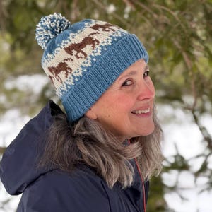 May include: A woman wearing a blue knit hat with a white pom pom and a brown and white patterned design. The hat features a repeating pattern of stylized horses.