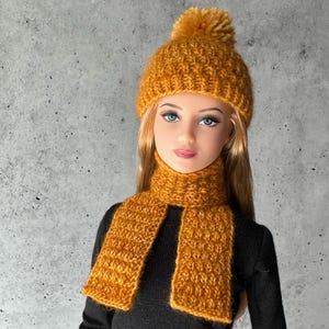 May include: A doll wearing a mustard yellow knitted beanie with a matching pom-pom on top and a coordinating mustard yellow knitted scarf. The doll has long blonde hair, blue eyes, and is dressed in a plain black long-sleeved shirt.