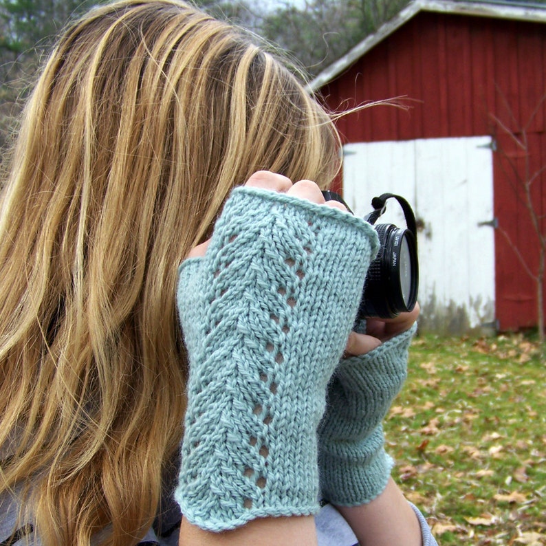 May include: A pair of light blue fingerless knitted gloves with a delicate lace pattern. The gloves are worn by a person holding a camera.