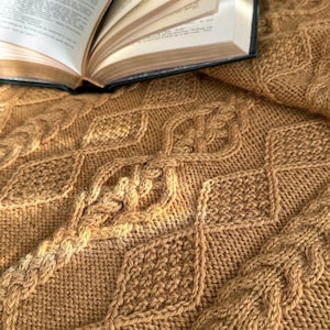 May include: A tan knitted blanket featuring a textured diamond pattern with intricate cable knit details. An open book with white pages and black printed text rests on the upper portion of the folded fabric, showcasing the detailed stitch work.