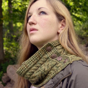 May include: A woman wearing a green and brown knitted cowl neck scarf with decorative buttons. The scarf is made of a chunky yarn and has a cable knit pattern.
