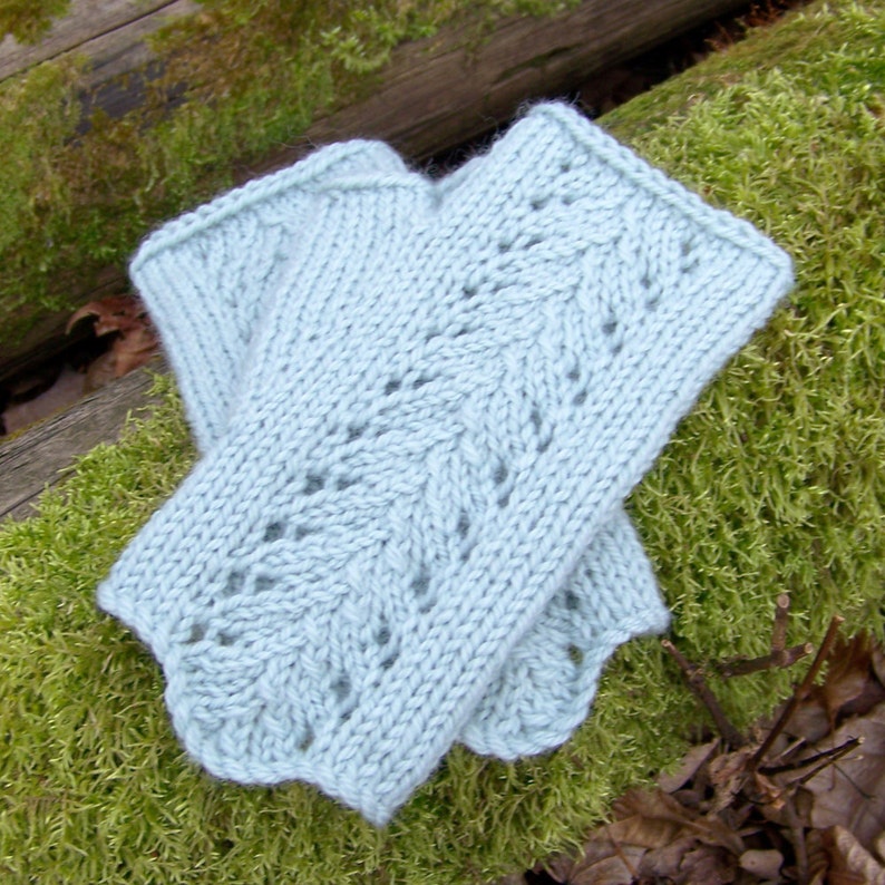 May include: A pair of light blue knitted fingerless gloves with a lacy pattern. The gloves are folded in half and are lying on a bed of green moss.