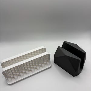 May include: Two geometric phone stands. One is white with a honeycomb pattern, and the other is black with a faceted design. Both are made of a solid material and are designed to hold a phone upright.