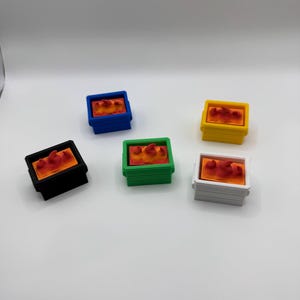May include: Six small, colorful, rectangular containers. Each container is a different color: black, blue, green, yellow, and white. The containers have an orange and red object inside.