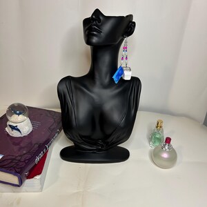 Puede incluir: Pendientes colgantes con un cubo blanco y un acento azul. Los pendientes están adornados con cuentas rosas y plateadas. Se exhiben en un busto negro. Otros elementos en la imagen incluyen una bola de nieve, libros y frascos de perfume.