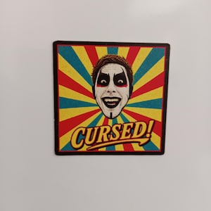 Peut inclure: Un aimant carr&eacute; pr&eacute;sentant une illustration graphique d'une personne avec du maquillage blanc, des yeux maquill&eacute;s de noir et un large sourire. Le fond se compose de rayons de soleil rouges, jaunes et bleu sarcelle. Le mot CURSED! est &eacute;crit en jaune.