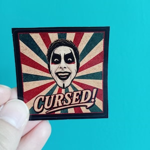 Peut inclure: Un autocollant carr&eacute; pr&eacute;sentant une illustration centrale d'une personne aux cheveux fonc&eacute;s avec un maquillage noir et rouge spectaculaire. Le fond se compose de rayons de soleil rouges et vert fonc&eacute;. Le mot CURSED! est imprim&eacute; en bas.