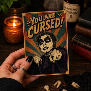 May include: A rectangular postcard featuring a vintage-style illustration of a person with dark makeup pointing forward. The text YOU ARE CURSED! is printed in bold yellow letters at the top against a background of teal and orange sunburst rays.