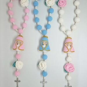 May include: Three handmade crochet rosaries in pink, blue, and white. Each rosary features a small Virgin Mary figure, a cross, and decorative flowers. The beads are crocheted in various colours, with a pearl-like string.