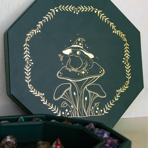 May include: A dark green octagonal dice tray and lid set. The lid features a gold-colored line art illustration of a frog wearing a wizard hat, sitting on a mushroom, surrounded by a delicate botanical wreath and small stars.