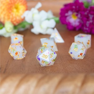 May include: A set of polyhedral dice made of translucent white resin with embedded colorful dried flowers and gold-colored numbers. The dice feature various geometric shapes, including a twenty-sided die prominently displaying the number 20.