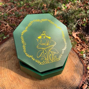 May include: A dark green octagonal box with a gold-colored line art illustration on the lid. The design features a frog wearing a pointed hat sitting on a large mushroom, surrounded by a circular wreath of leaves and small stars.