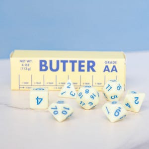 May include: A still life featuring a stick of butter and several dice. The butter package is yellow with blue text that reads "BUTTER" and "GRADE AA". The dice are off-white with blue numbers.