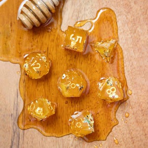 May include: A set of seven translucent amber-colored polyhedral dice featuring embedded gold flakes and small bee motifs, with white numbering. A wooden honey dipper rests in a pool of golden honey surrounding the dice on a wooden surface.