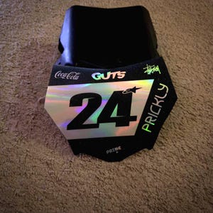May include: Black motocross number plate with the number 24 in large, bold, black font. The plate has a holographic silver and iridescent finish. The words "GUTS", "PRICKLY", and "PRIME" are visible, along with the Coca-Cola and Stussy logos.