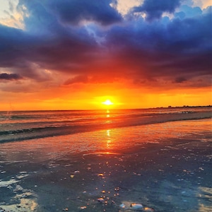 Könnte beinhalten: Ein lebendiger Sonnenuntergang über dem Meer. Der Himmel wechselt von tiefem Blau zu feurigem Orange und Rot, wobei sich die Sonne auf dem Wasser und dem nassen Sand spiegelt. Am Horizont ist eine entfernte Küstenlinie zu sehen.