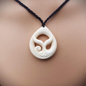 Pode incluir: Colar com pingente creme com design de cauda de baleia. O pingente em forma de gota pende de um cordão preto. O design da cauda de baleia é esculpido no pingente, com detalhes intrincados. É uma joia.