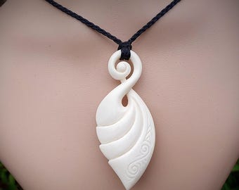 May include: Cream-colored pendant necklace with a carved, stylized design. The pendant is suspended from a black cord. The pendant features a spiral at the top and a leaf-like shape with carved details.