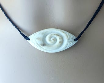 May include: A light blue oval pendant necklace with a spiral design and black cord. The pendant has a carved spiral in the center and leaf-like details on the sides. The necklace is displayed against a neutral background.