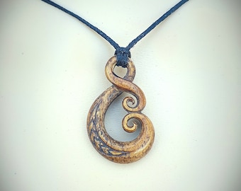 May include: A wooden pendant necklace with a swirling, double-spiral design. The pendant is a light brown color with darker brown etched details. It hangs from a dark blue cord, creating a simple, natural aesthetic.