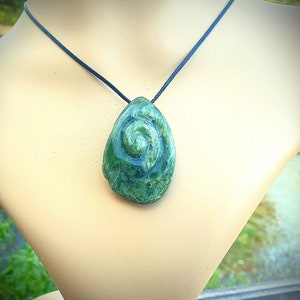 May include: A green stone pendant necklace with a spiral design. The pendant is teardrop-shaped and hangs from a black cord. The stone has a textured surface and varying shades of green.