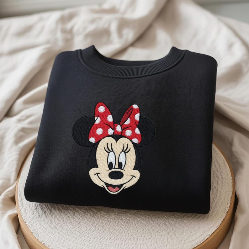 May include: Black sweatshirt featuring an embroidered Minnie Mouse design. The design includes Minnie's face, a red bow with white polka dots, and black ears. The sweatshirt is folded neatly on a light-colored surface.