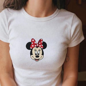 May include: White t-shirt featuring a cartoon image of Minnie Mouse. The design includes Minnie's face with black ears, a red bow with white polka dots, and a smiling expression. The shirt has short sleeves and a crew neck.