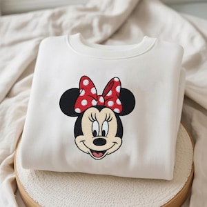 May include: A white sweatshirt featuring an embroidered Minnie Mouse design. The design includes Minnie's face, black ears, and a red bow with white polka dots. The sweatshirt is folded and placed on a wooden surface.