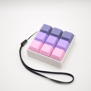 May include: A small, white rectangular device with nine square buttons in shades of pink, purple, and blue. A black lanyard is attached to the device. The buttons are arranged in three rows of three.