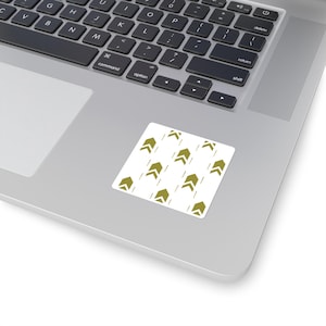 May include: A white square sticker with a repeating pattern of olive green upward-pointing arrows and vertical lines. The sticker is placed on a silver laptop.
