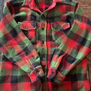 May include: A button-down flannel shirt in a classic plaid pattern of red, green, black, and brown. The shirt has two chest pockets and buttoned cuffs. The collar is red. The brand name is visible on the collar.